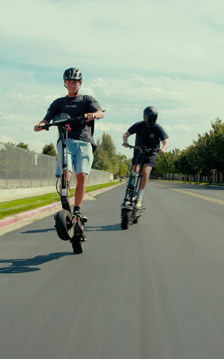 Two people riding Teewing electric scooters on a road with trees and a fence in the background.